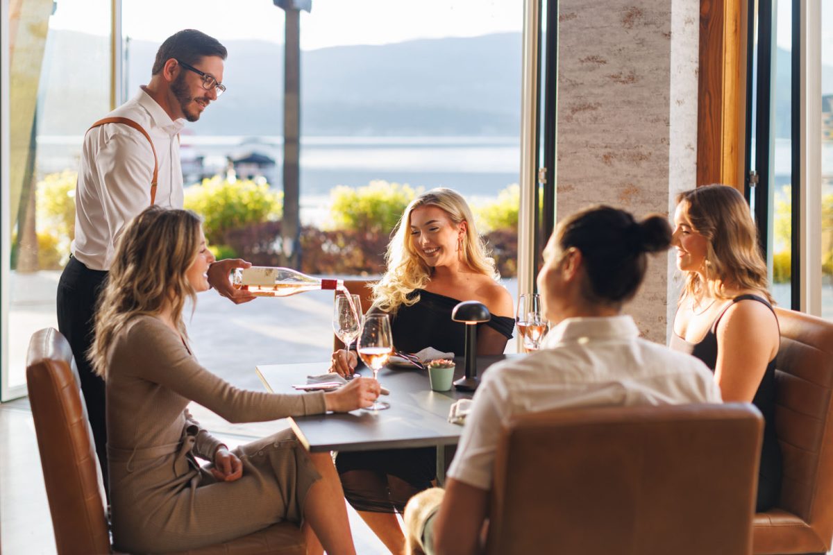 Maestro's restaurant interior in Kelowna of guests being poured wine and laughing and smiling with Okanagan Lake in background