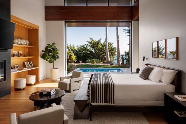 Interior of luxury primary bedroom looking out open doors to a swimming pool and palm trees overlooking the ocean